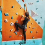 Young,Woman,Climbing,Up,On,Practice,Wall,In,Gym,,Rear
